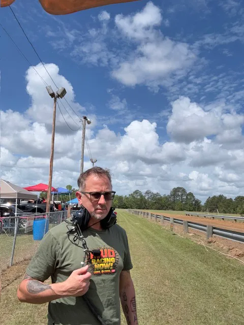 Sergio Myers carrying field audio equipment beside an outdoor racing course